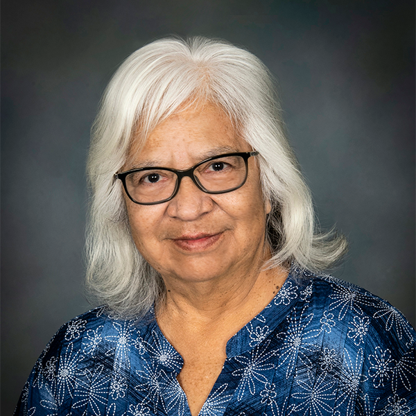 Headshot of Carol Cornelius in front of a grey backdrop. She has light colored hair and is wearing black framed glasses and a blue blouse